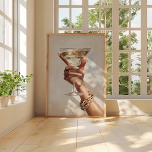Op de afbeelding: Een ingelijste prent van een hand die een martiniglas vasthoudt. De hand is versierd met ringen en een gouden armband. Het martiniglas is gevuld met een heldere vloeistof. Het kunstwerk wordt tentoongesteld in een kamer met natuurlijk licht.