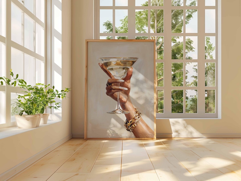 Op de afbeelding: Een ingelijste prent van een hand die een martiniglas vasthoudt. De hand is versierd met ringen en een gouden armband. Het martiniglas is gevuld met een heldere vloeistof. Het kunstwerk wordt tentoongesteld in een kamer met natuurlijk licht.