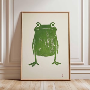 May include: A green frog illustration printed on a white background in a wooden frame. The frog has large, round eyes and a simple, stylized body.