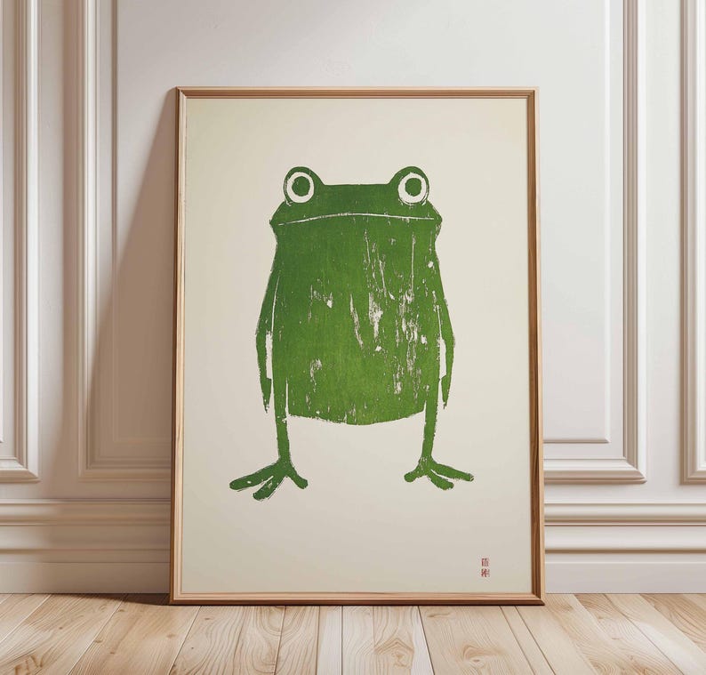 May include: A green frog illustration printed on a white background in a wooden frame. The frog has large, round eyes and a simple, stylized body.