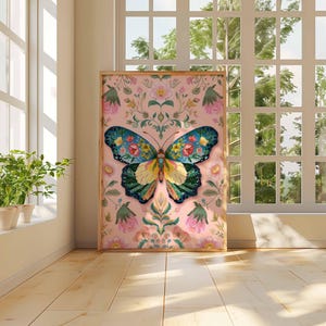 May include: A framed art print featuring a colorful butterfly design. The butterfly has blue, green, yellow, and floral details. The background is a soft pink with floral and botanical elements. The artwork is displayed in a room with natural light.