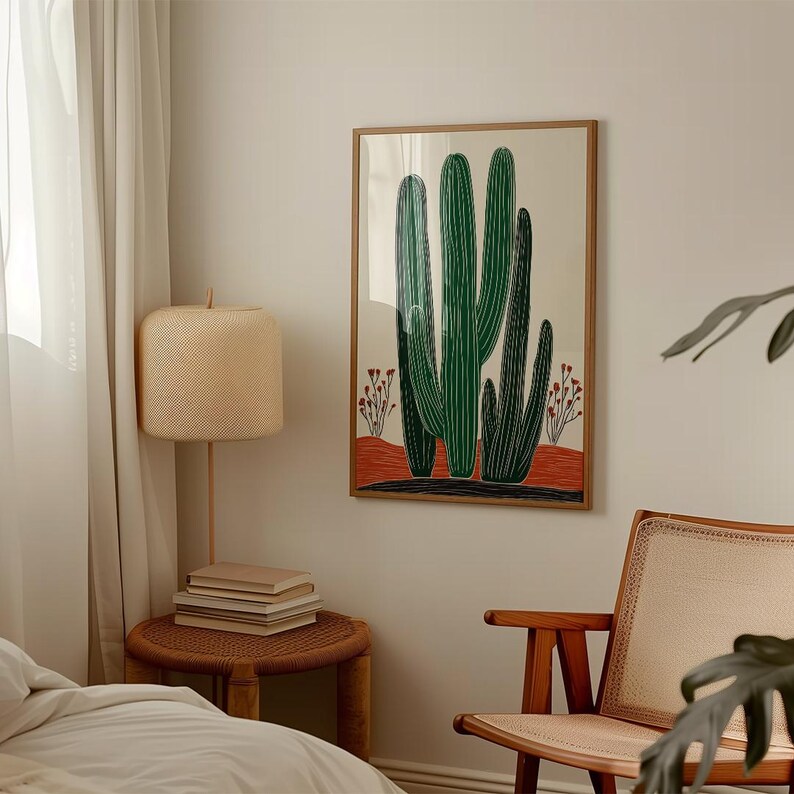 May include: A framed art print featuring a stylized illustration of green cacti against a beige background. The artwork includes a red and black base, with a wooden frame. The print is displayed in a well-lit room, alongside a woven side table and a chair.