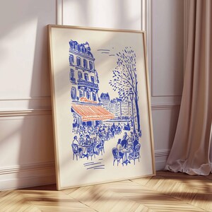May include: A framed art print depicts a Parisian cafe scene in blue and orange. The illustration shows a building with an orange awning, tables, and chairs, with people seated at them. A tree and other buildings are in the background.