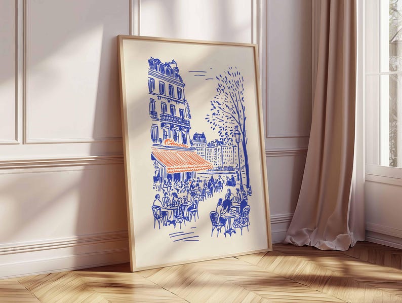May include: A framed art print depicts a Parisian cafe scene in blue and orange. The illustration shows a building with an orange awning, tables, and chairs, with people seated at them. A tree and other buildings are in the background.