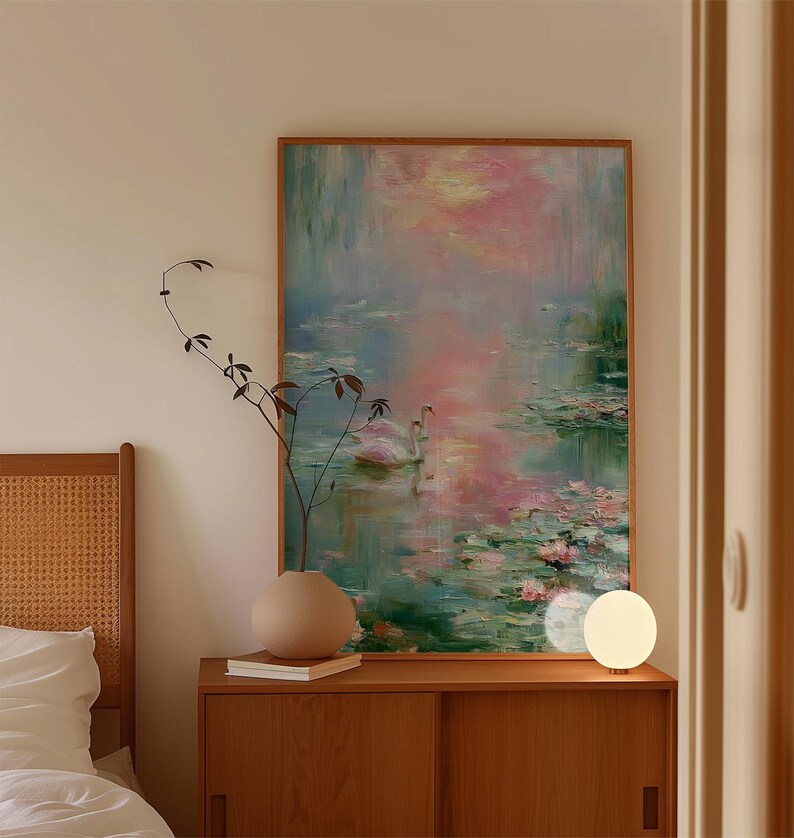 May include: A framed painting depicts a serene water scene with pink and blue hues, featuring swans and lily pads. The artwork hangs above a wooden cabinet with a vase and a lamp. The room has a neutral color palette.