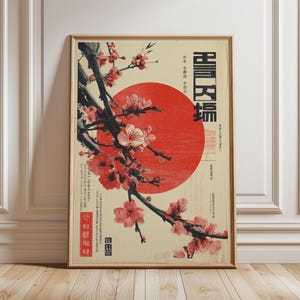 Op de afbeelding: Ingelijste kunstprint met een Japans geïnspireerd ontwerp. Een rode zon op de achtergrond, met een tak met kersenbloesems op de voorgrond. De print heeft Japanse tekst en zit in een houten lijst.