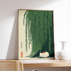 May include: Framed art print featuring a serene scene of a white and orange cat resting under a weeping willow tree. The artwork has a Japanese aesthetic, with green foliage and a beige background. The frame is a light brown color.