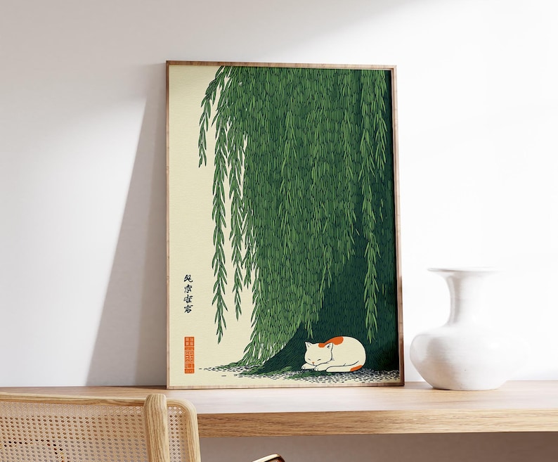 May include: Framed art print featuring a serene scene of a white and orange cat resting under a weeping willow tree. The artwork has a Japanese aesthetic, with green foliage and a beige background. The frame is a light brown color.