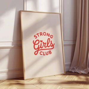 Pu&ograve; includere: Una stampa incorniciata con le parole "STRONG Girls CLUB" in rosso e un design a cuore. La stampa &egrave; in una cornice di legno chiaro e si appoggia a un muro bianco. Una tenda beige e una finestra sono visibili sullo sfondo.