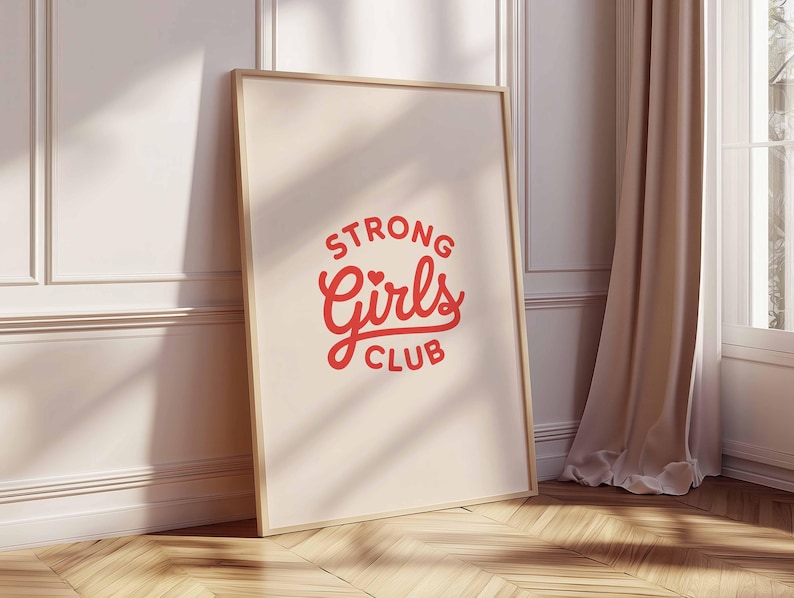 Pu&ograve; includere: Una stampa incorniciata con le parole "STRONG Girls CLUB" in rosso e un design a cuore. La stampa &egrave; in una cornice di legno chiaro e si appoggia a un muro bianco. Una tenda beige e una finestra sono visibili sullo sfondo.