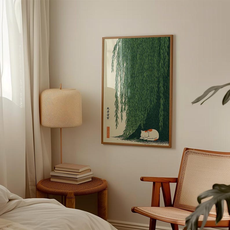 May include: A framed print depicts a green weeping willow tree with a white and orange cat sleeping beneath its branches. The artwork has a wooden frame and is displayed on a neutral-colored wall. A woven lamp and side table are in the foreground.