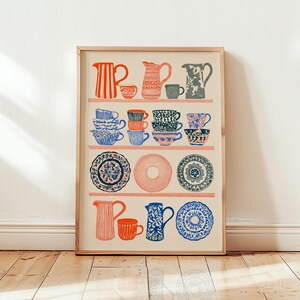 May include: A framed art print featuring a collection of illustrated kitchenware arranged on shelves. The print displays various pitchers, teacups, and plates in shades of red, blue, and gray against a cream background, organized in rows.