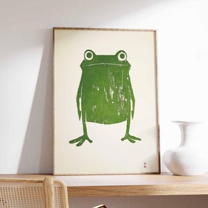May include: A framed print of a green frog with a white background. The frog is stylized and has a simple design.