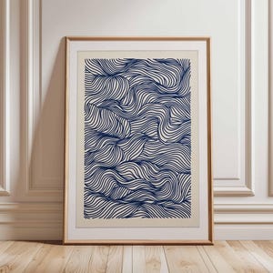 Puede incluir: Impresión artística enmarcada con un patrón de ondas azul marino sobre un fondo blanquecino. La obra de arte se presenta en un marco de madera marrón claro, apoyada contra una pared blanca y un suelo de madera.