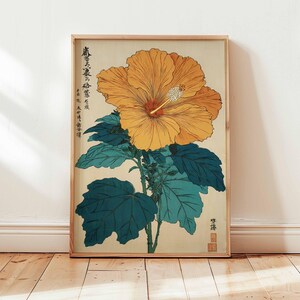 May include: A framed botanical print depicting a large, yellow hibiscus flower with a red centre and white stamen. The flower is set against a cream background with teal leaves and stems. The print has Japanese text and is in a wooden frame.