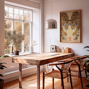 May include: A sunlit room with a wooden desk and two chairs. A framed artwork of two tigers is on the wall. A large window offers a view of trees. A silver pendant light hangs above the desk. Plants and decorative items are on the desk and windowsill.