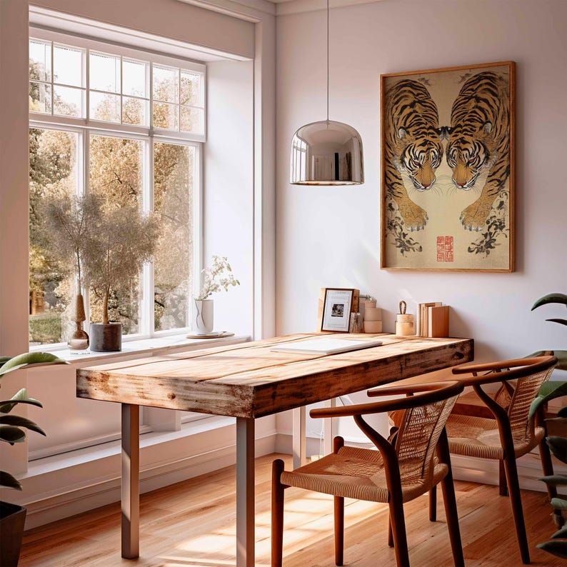 May include: A sunlit room with a wooden desk and two chairs. A framed artwork of two tigers is on the wall. A large window offers a view of trees. A silver pendant light hangs above the desk. Plants and decorative items are on the desk and windowsill.