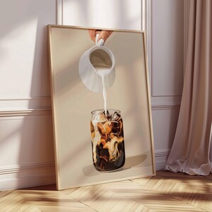 May include: A framed print depicts a refreshing iced coffee. A white pitcher pours milk into a glass filled with ice and dark coffee. The artwork is set against a neutral background, leaning against a wall with a wooden floor.