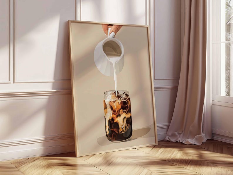 May include: A framed print depicts a refreshing iced coffee. A white pitcher pours milk into a glass filled with ice and dark coffee. The artwork is set against a neutral background, leaning against a wall with a wooden floor.
