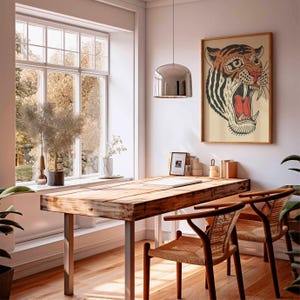 May include: A wooden desk with metal legs and two woven chairs sits in a bright room. A framed tiger print hangs on the wall. A large window offers a view of trees. A silver pendant light hangs above the desk.