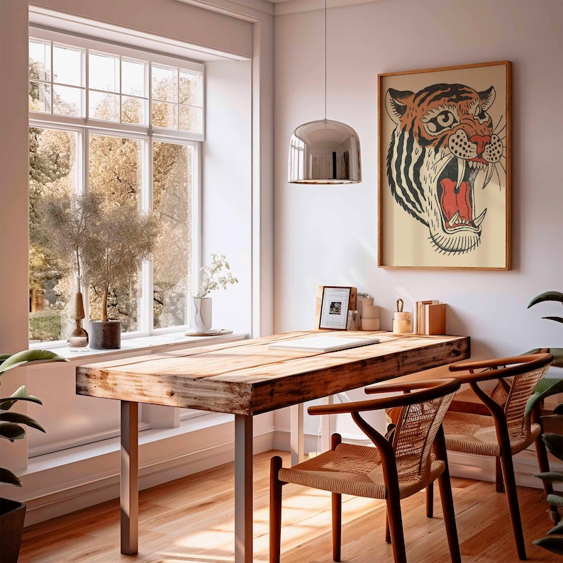 May include: A wooden desk with metal legs and two woven chairs sits in a bright room. A framed tiger print hangs on the wall. A large window offers a view of trees. A silver pendant light hangs above the desk.