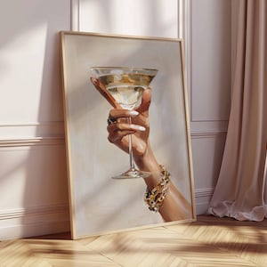 Op de afbeelding: Een ingelijste kunstdruk toont een hand die een martini-glas vasthoudt. Het glas bevat een heldere vloeistof en heeft een gouden rand. De hand draagt een gouden armband en ringen, en de nagels zijn wit geschilderd.