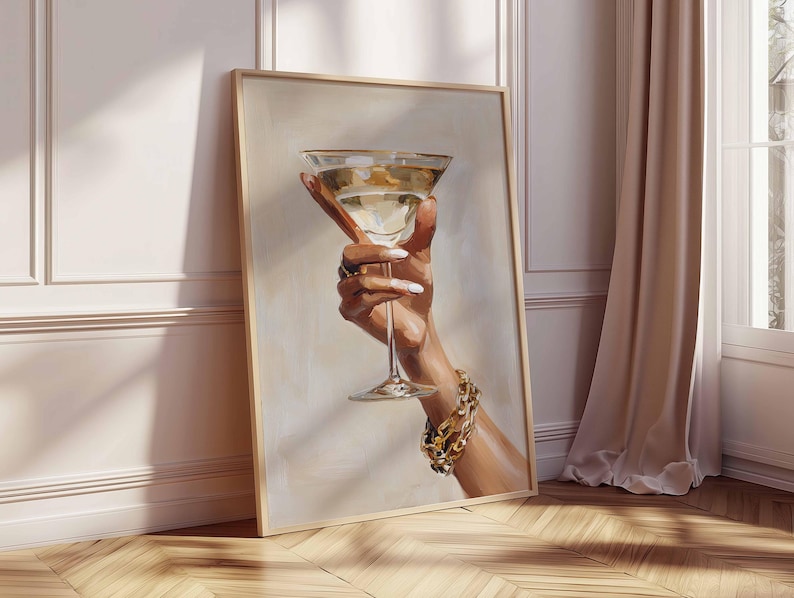 Op de afbeelding: Een ingelijste kunstdruk toont een hand die een martini-glas vasthoudt. Het glas bevat een heldere vloeistof en heeft een gouden rand. De hand draagt een gouden armband en ringen, en de nagels zijn wit geschilderd.