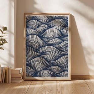 May include: A framed art print featuring a navy blue and cream-colored wave pattern. The artwork is displayed in a light-colored wooden frame and leans against a wall. The wave design is composed of numerous parallel lines.