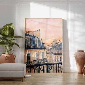 May include: A framed watercolour print depicting Parisian rooftops with blue-grey mansard roofs and balconies, set against a pink and orange sunset. The artwork is in a light wood frame and leans against a white wall.