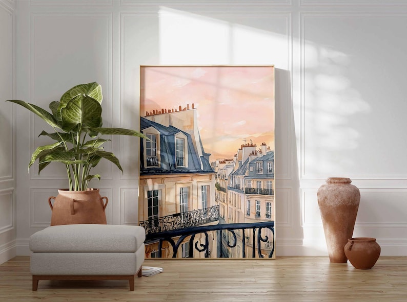 May include: A framed watercolour print depicting Parisian rooftops with blue-grey mansard roofs and balconies, set against a pink and orange sunset. The artwork is in a light wood frame and leans against a white wall.