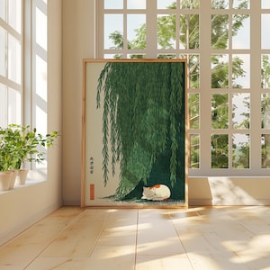 May include: A framed print featuring a green weeping willow tree with a sleeping white and orange cat beneath its branches. The artwork is in a room with a window and plants.