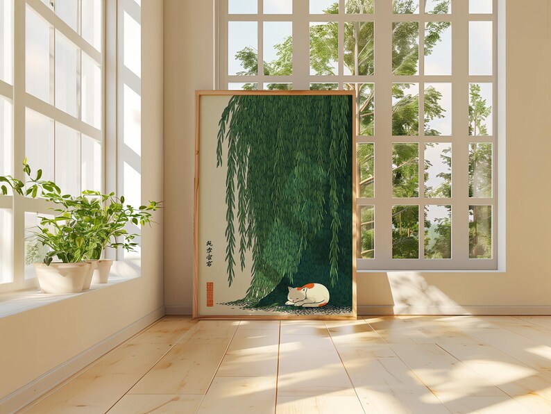 May include: A framed print featuring a green weeping willow tree with a sleeping white and orange cat beneath its branches. The artwork is in a room with a window and plants.