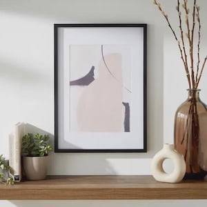 May include: A framed abstract art print in a black frame. The artwork displays soft pink, grey, and black shapes with a thin black line. A brown glass vase with branches, a small white vase, and a potted plant are on a wooden shelf.