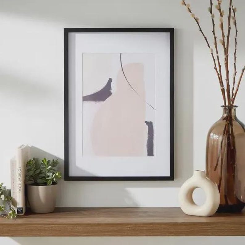 May include: A framed abstract art print in a black frame. The artwork displays soft pink, grey, and black shapes with a thin black line. A brown glass vase with branches, a small white vase, and a potted plant are on a wooden shelf.