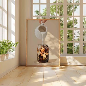 May include: A framed print depicts a refreshing iced coffee. A hand pours milk from a white pitcher into a glass filled with ice and dark coffee. The artwork is set against a neutral background, with natural light from a window.