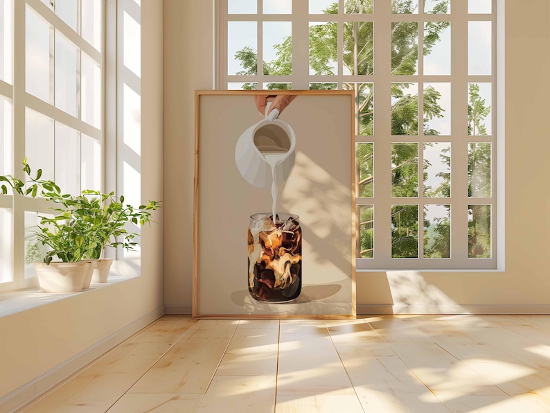 May include: A framed print depicts a refreshing iced coffee. A hand pours milk from a white pitcher into a glass filled with ice and dark coffee. The artwork is set against a neutral background, with natural light from a window.