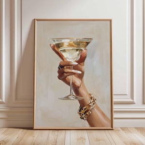Op de afbeelding: Een ingelijste kunstdruk toont een hand die een martiniglas vasthoudt gevuld met een gouden vloeistof. De hand is versierd met ringen en een gouden schakelarmband. De achtergrond is neutraal beige en de lijst is lichtbruin.