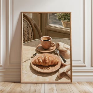 May include: A framed print depicting a breakfast setting. A croissant is on a plate beside a coffee cup on a table. A spoon, fork, and napkin are also present. The scene is illuminated by warm light, creating a cosy atmosphere.