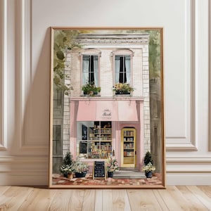 Può includere: Una stampa ad acquerello incorniciata di una libreria pittoresca con una tenda rosa e una porta gialla. La facciata del negozio è in mattoni bianchi, con due finestre sopra la tenda. La stampa include il testo "Book Retreive".