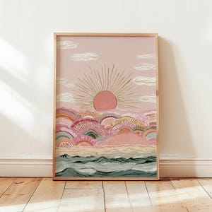 以下が含まれることがあります： ピンクの太陽が黄金の光を放ち、ピンク、ピーチ、セージグリーンの半円形の丘が重なる風景を描いた額装アートプリント。丘の下には砂浜が広がり、濃いティールブルーの波が打ち寄せる様子が描かれています。