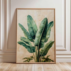 May include: A framed botanical print depicting a cluster of large, green banana leaves against a beige backdrop. The artwork is enclosed in a light brown wooden frame, creating a natural and elegant aesthetic. The painting is leaning against a white wall and wooden floor.