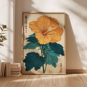 May include: A framed botanical print depicting a large orange hibiscus flower with a white and red centre. The flower is set against a beige background with dark teal leaves and stems. Japanese text is visible.