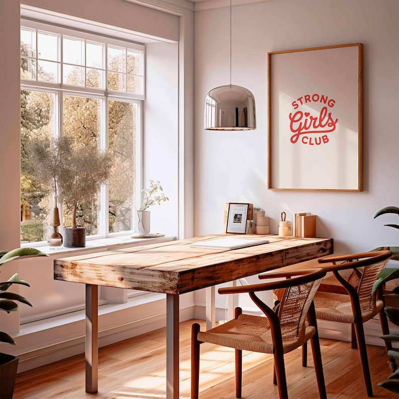 Pu&ograve; includere: Un ufficio domestico luminoso e arioso con una grande scrivania in legno e due sedie intrecciate. Una stampa incorniciata sulla parete recita "STRONG Girls CLUB" in rosso. Una grande finestra offre una vista sugli alberi e sulla luce naturale.