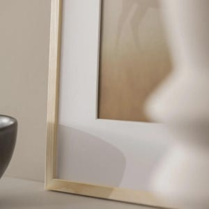 May include: A framed picture with a light wood frame and a white mount. The artwork has a blurred image with tan and brown tones. A dark grey bowl is visible in the lower left corner. The frame is made of light-coloured wood.