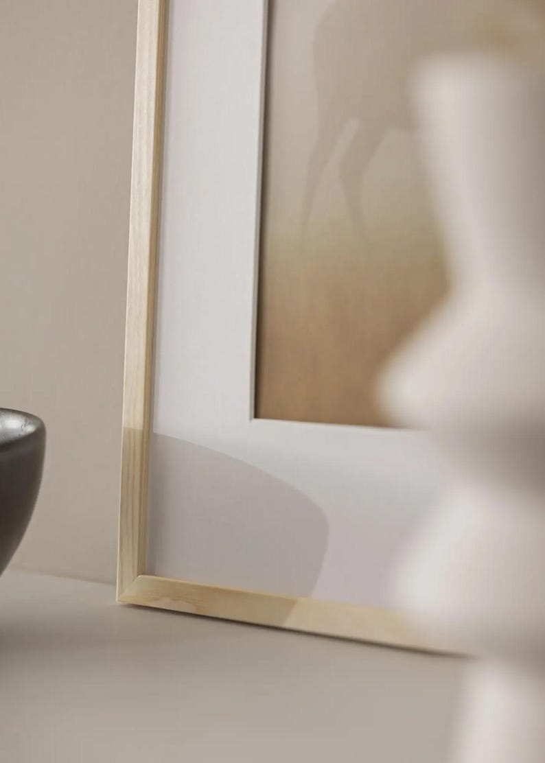 May include: A framed picture with a light wood frame and a white mount. The artwork has a blurred image with tan and brown tones. A dark grey bowl is visible in the lower left corner. The frame is made of light-coloured wood.