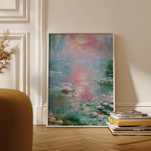 May include: Framed artwork depicting a serene lake scene with two swans. The painting features a soft color palette of pink, blue, and green, with lily pads and reflections in the water. The artwork is leaning against a white wall.