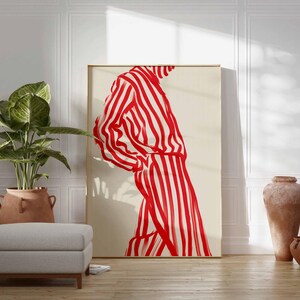 Puede incluir: Una gran impresi&oacute;n art&iacute;stica enmarcada que presenta una figura con una prenda a rayas rojas y blancas. La obra de arte se exhibe en una habitaci&oacute;n con una planta en maceta, un otom&aacute;n gris y cer&aacute;mica decorativa. La habitaci&oacute;n tiene un ambiente luminoso y aireado.