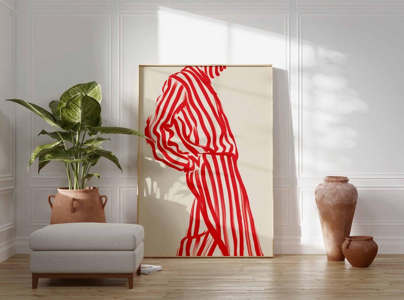 Puede incluir: Una gran impresi&oacute;n art&iacute;stica enmarcada que presenta una figura con una prenda a rayas rojas y blancas. La obra de arte se exhibe en una habitaci&oacute;n con una planta en maceta, un otom&aacute;n gris y cer&aacute;mica decorativa. La habitaci&oacute;n tiene un ambiente luminoso y aireado.