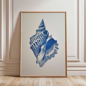 Könnte beinhalten: Gerahmter Druck mit einer detaillierten blauen Muscheldarstellung auf neutralem Hintergrund. Das Kunstwerk wird in einem hellen Holzrahmen präsentiert, der vor einer weißen Wand und einem Holzboden steht, was ein maritimes Thema hervorruft.