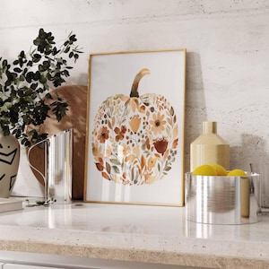 May include: A framed watercolor print of a pumpkin composed of autumn-colored flowers and leaves. The artwork has a gold frame and is displayed on a white countertop. Other decorative items include a vase, a pitcher, and a bowl with lemons.
